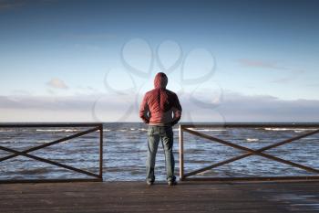 Young man in hood looking on the sea from wooden pier at morning. Rear view photo