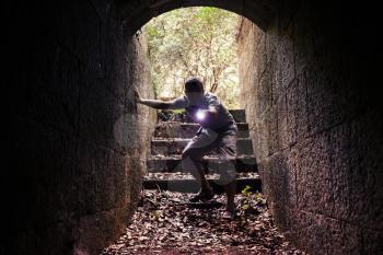 Young man with a flashlight enters dark stone tunnel and looks inside with attention and fear