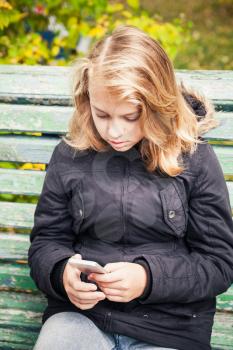 Beautiful Caucasian blond teenage girl in black jacket sitting on green bench with smartphone in hands