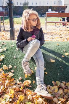 Beautiful blond teenage girl in glasses sitting in autumnal European city park with falling leaves