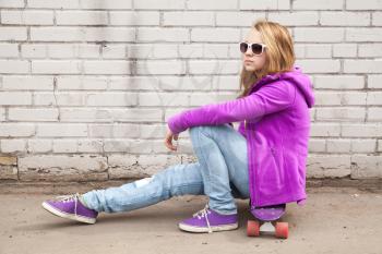 Beautiful blond teenage girl in sunglasses sits on skateboard near gray brick wall