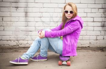 Beautiful blond teenage girl in sunglasses sits on skateboard near white brick wall
