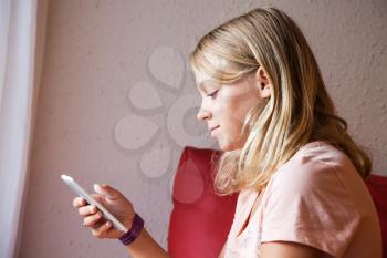 Cute Caucasian blond teenage girl in pink t-shirt using smartphone for messaging, indoor closeup profile portrait