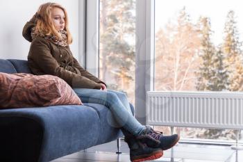 Beautiful blond Caucasian teenage girl in warm clothes sitting on sofa near the window in hallway