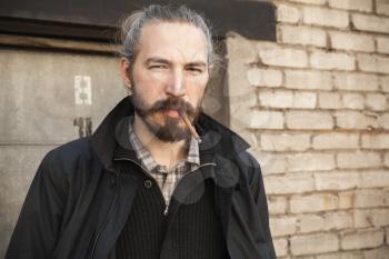 Bearded man smoking cigarette over old brick wall, outdoor portrait with selective focus