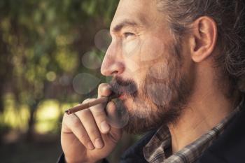 Bearded Asian man smoking cigar, outdoor profile portrait with selective focus