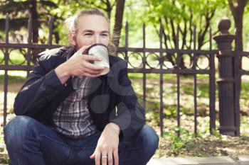 Bearded Asian man sitting on the sidewalk in park and drinking coffee from paper cup
