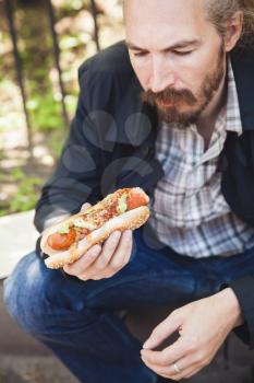 Bearded Asian man eating hot dog in summer park, outdoor portrait with selective focus