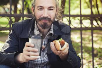 Bearded Asian man eating hot dog with coffee in summer park, outdoor portrait with selective focus