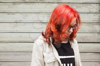 Sad Caucasian teenager girl in glasses with bright red hair, closeup outdoor portrait over green grungy wooden wall