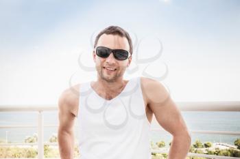 Young sporty smiling Caucasian man in white shirt and black sunglasses. Outdoor summer portrait with sea on a background