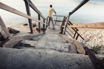 Young man stands on old wooden stairway going down to the sea coast
