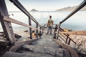 Young man stands on old wooden stairway going down to the sea coast in morning 