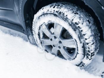 Car wheel with studded tire standing on winter road with deep snow, blue toned photo with selective focus