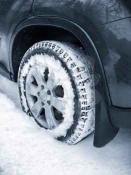 Modern car wheel with studded tire standing on winter road with deep snow, blue toned vertical photo with selective focus