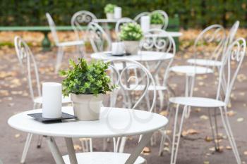 Outdoor cafeteria background interior, metal white chairs and tables with decorative green plants in pots and candles 