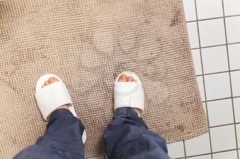 Male feet in white slippers stand on brown carpet in bathroom