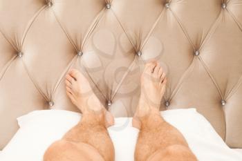 Relaxing male feet lay on white pillow near soft headboard in bedroom, luxury rest lifestyle