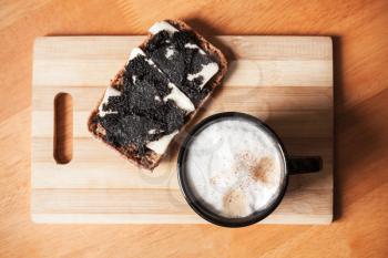 Cup of cappuccino and a sandwich with black caviar stand on wooden table, top view, closeup photo with selective focus