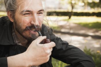 Young smiling asian bearded man smoking pipe in summer park, close up portrait