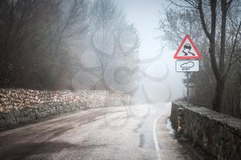 Slippery road in rainy weather, warning roadsign stands on mountain roadside, rural landscape