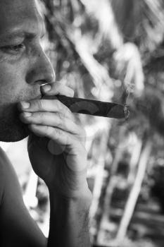 Young European man smokes big cigar, close up vertical monochrome photo with selective focus. Dominican Republic