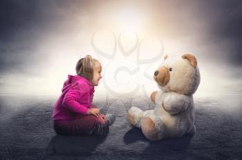 Small cute girl sits and looks at toy bear on grey background