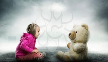 Small cute girl sits and looks at toy bear on grey and white background