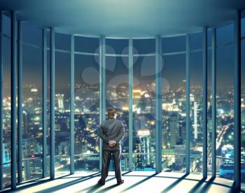 Businessman is standing in front of night view of buildings from the last floor of high office building