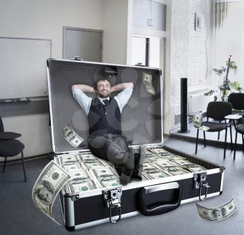 Happy businessman sits in the case full of dollars in the office