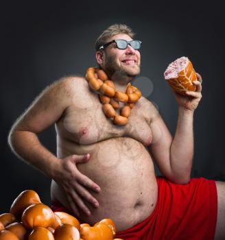 Happy man in glasses with sausages round his neck holds a big wurst in his hand over grey
