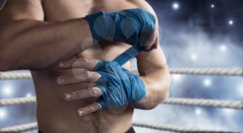 Close-up of hand of strength boxer who pulls bandage before the fight or training. Boxing ring on the background.