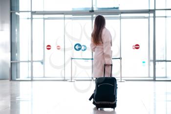 Young woman with a suitcase goes to exit the station. Glass doors on the background. Suitable for bus, railway, metro station or airport. 