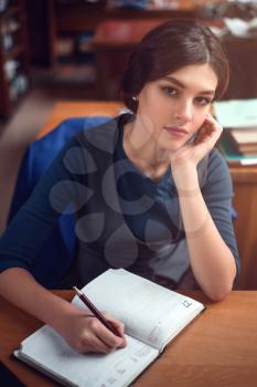 Young woman sitting at the table and writing in her notebook. Library reading room on the background