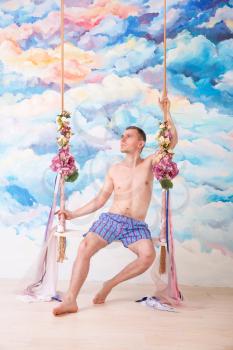Amorous man in pants sitting on wooden swing with ropes agaist abstract watercolor background. Romantic wedding concept.