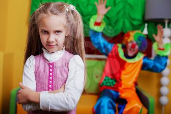 Upset little girl standing in the playroom.  Funny clown on the background.