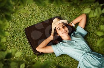 Young woman in light blue dress and white hat put her head on suitcase and dreams of a future journey. Girl lies on green grass. Travel waiting concept.
