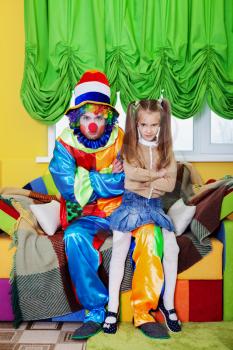 Funny clown with the little girl sit on a sofa. Celebrating birthday party 