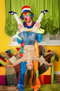 Amusing clown holds the little girl head over heels. Colourful couch on the background. 