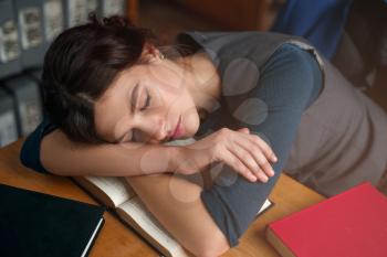 Young girl fell asleep while reading a book in college library. Bookshelf on the background. Student examination concept.