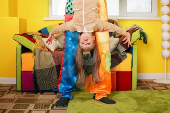 Amusing clown holds the little girl head over heels. Colourful couch on the background.
