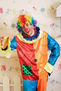 Serious clown in funny costume standing in the playroom. Decorative birds and nesting box on the background.