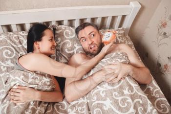 Young woman show alarm clock to her husband in the bed.