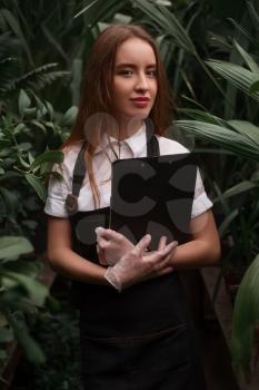 Young florist in apron holds clipboard in hands against greenhouse with exotic plants.