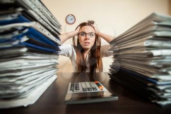 Doomed female accountant against big stacks of documents and calculator