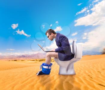 Man in glasses with laptop sitting on the toilet bowl in sandy desert