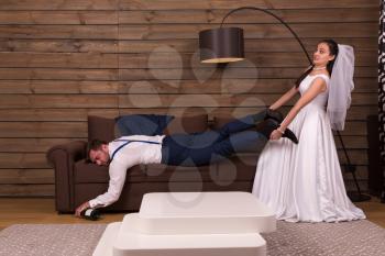 Bride is trying to wake up a drunk sleeping on couch groom. After wedding celebration. Wooden interior of the room on background