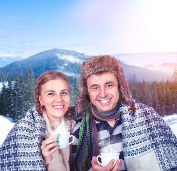 Love couple wrapped in warm plaid with cups of hot beverage in hands against snowy mountains and pine forest on background
