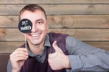Young man holds funny icon on a stick with  just married inscription and show thumbs up sign, wooden background. Fun photo props and accessories for photo shoots