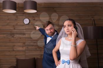 Portrait of bride talking by phone and dissatisfied groom showing on watch, wooden room on background.